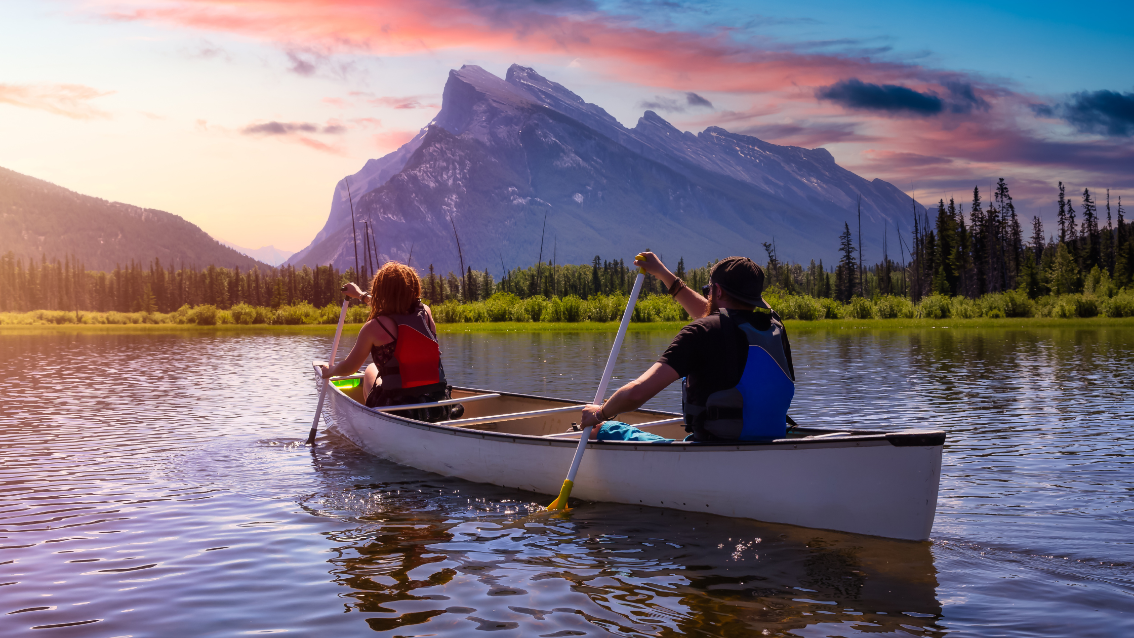 Man and woman rowing a canoe on a lake with sunset and mountains in the background