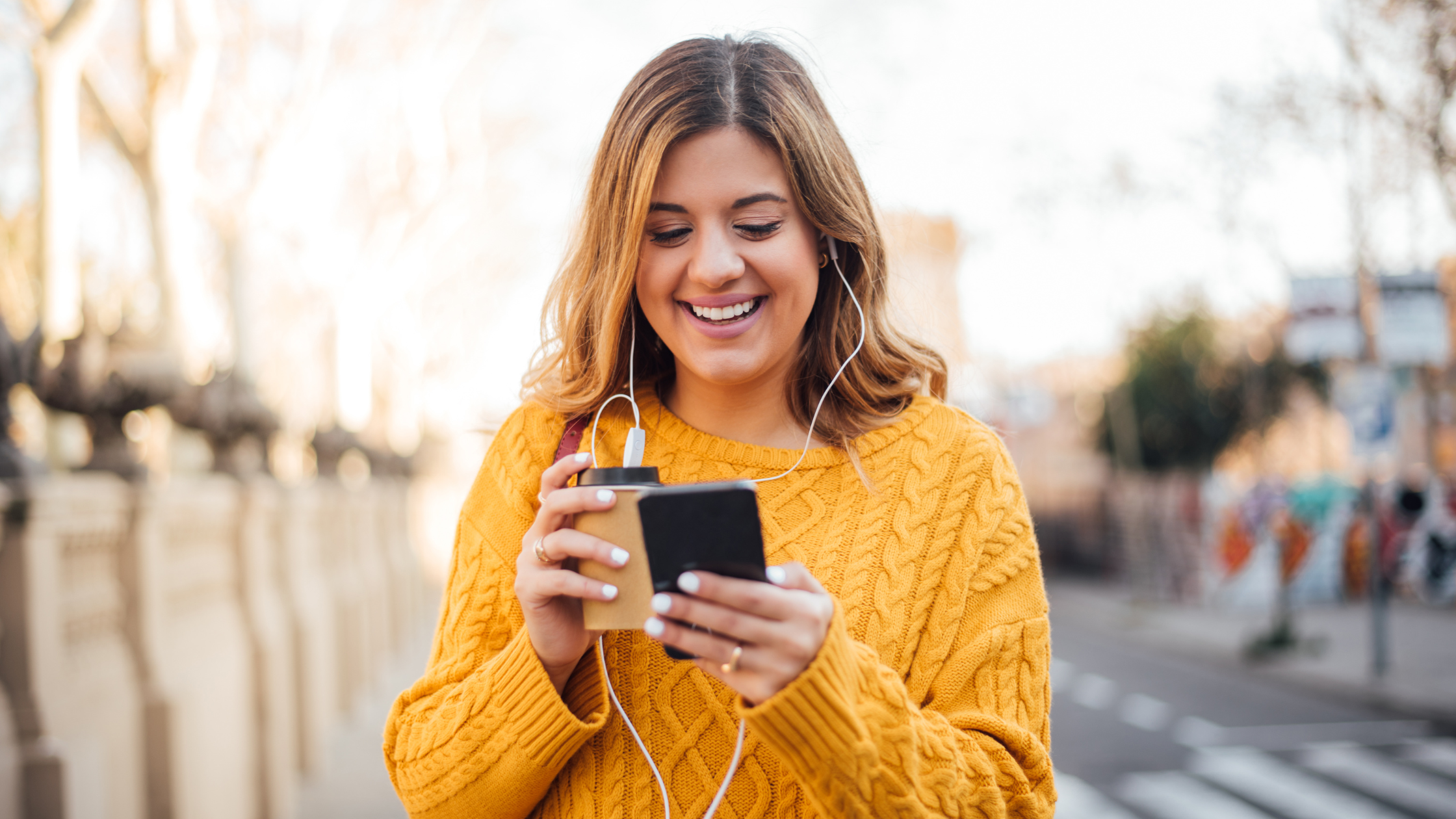 Woman in a yellow sweater, outside, smiling down at her cell phone